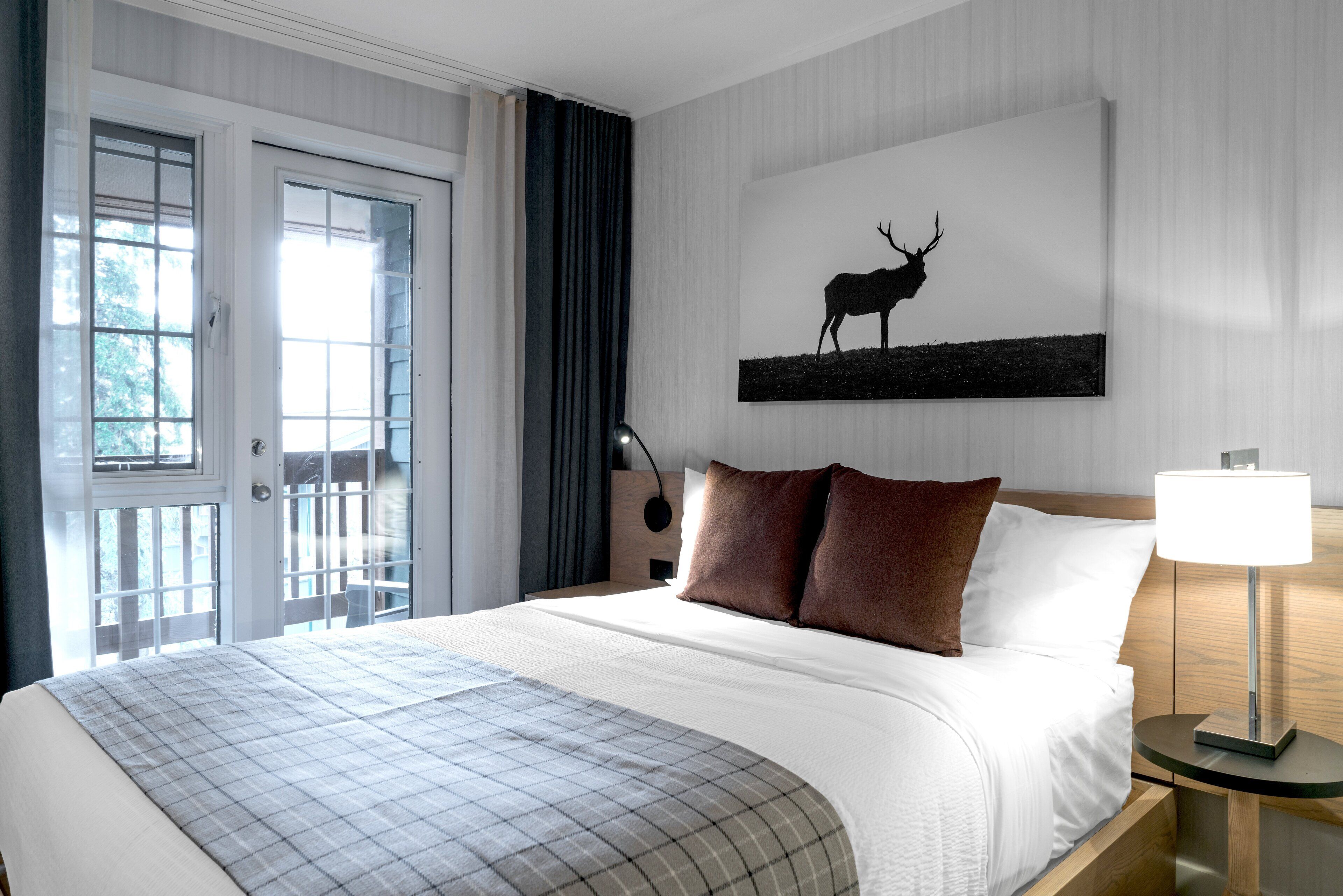 Une chambre d’hôtel avec un lit, une table de chevet avec une lampe, un tableau représentant un cerf et une porte donnant sur un balcon.