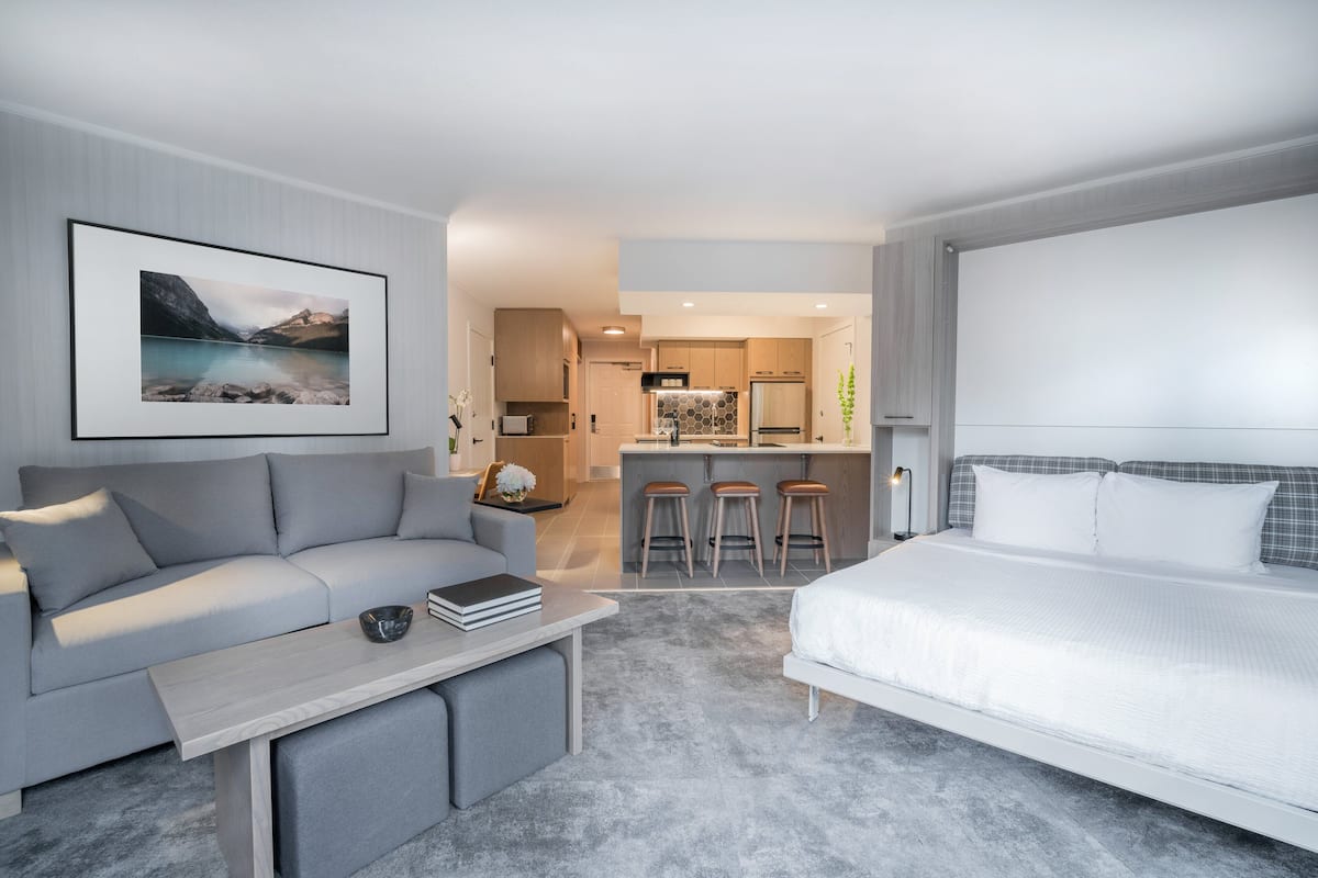 A modern hotel room with a grey sofa, a small coffee table, a bed with white bedding, and a kitchen area in the background.