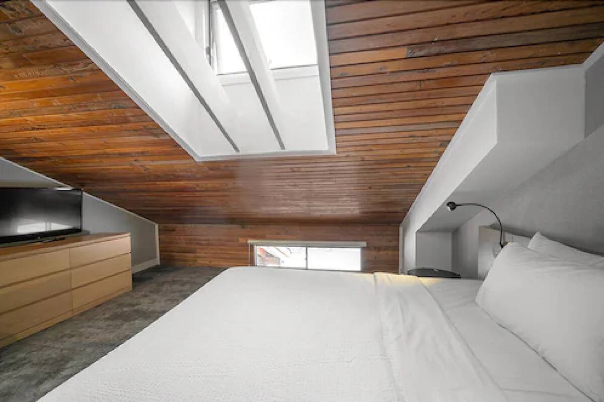 A bedroom with a sloped ceiling, wooden paneling, a bed, a TV, and a small window.