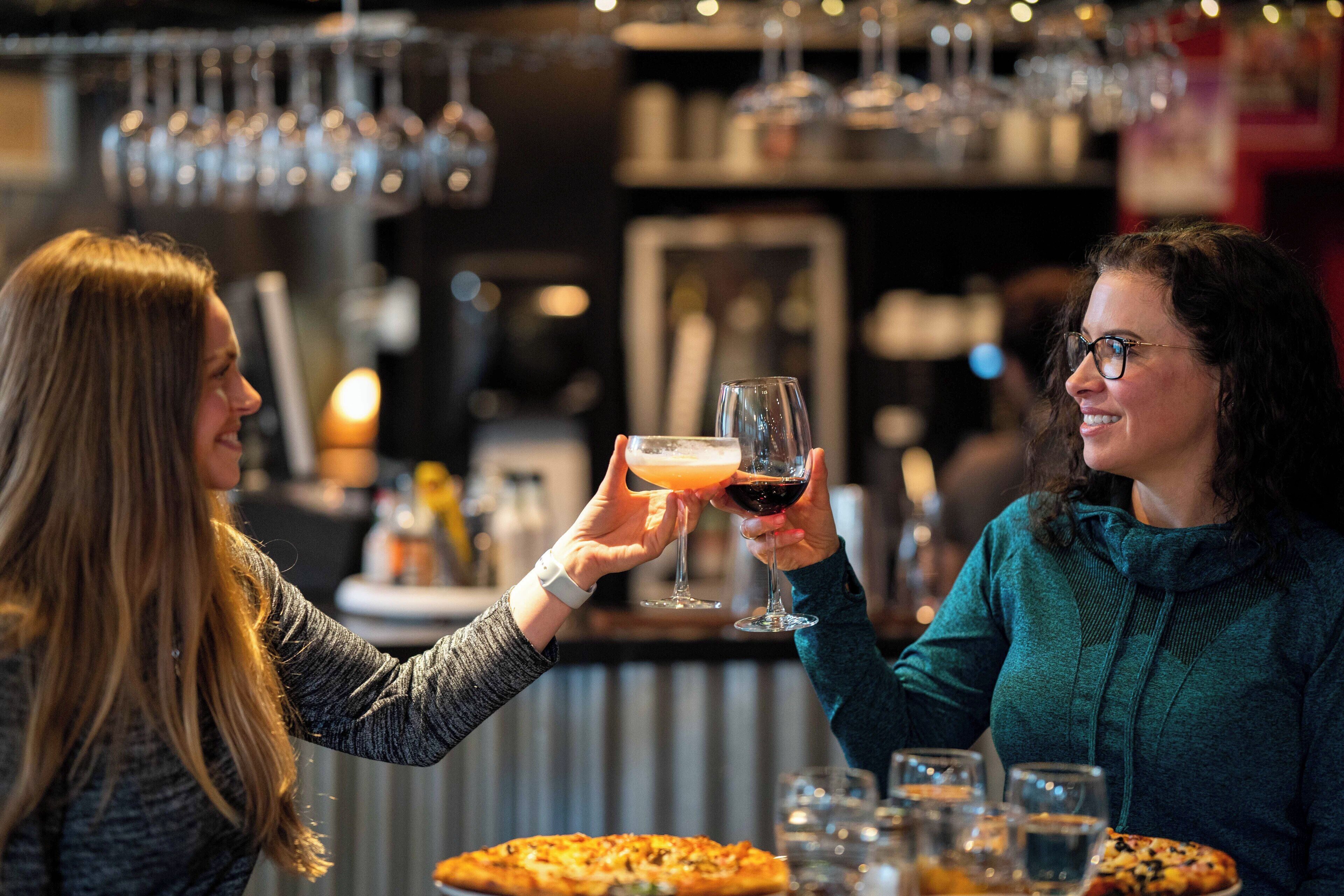 Dos personas brindando con bebidas en un bar, con una pizza sobre la mesa.