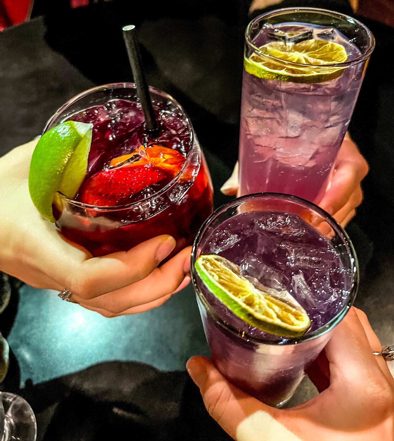 Three glasses of beverages with lime slices and a strawberry, held by hands.