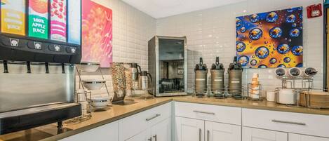 Una cocina moderna con cafetera, hervidor y prensa francesa. Hay un cuadro grande en la pared y una máquina expendedora con opciones de zumo de fruta.