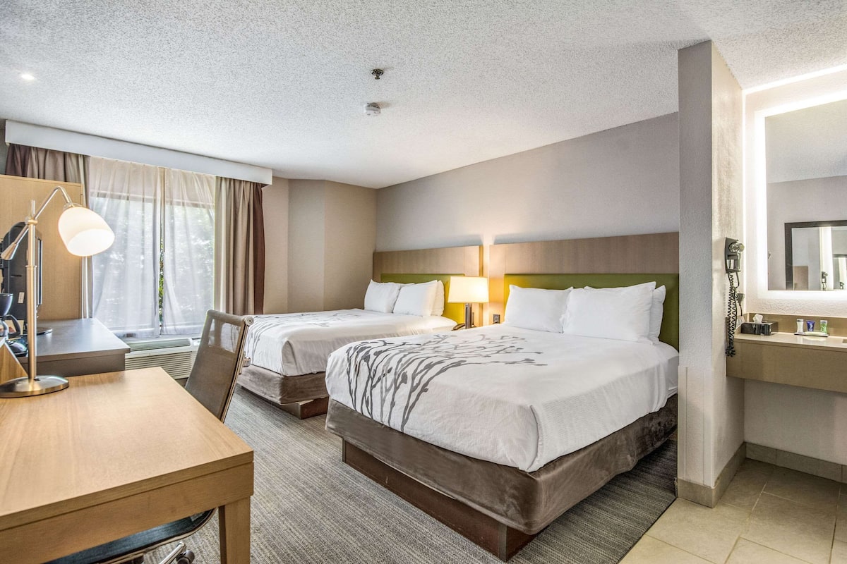 Ein Hotelzimmer mit zwei Betten, einem Schreibtisch, einem Stuhl, einer Lampe, einem Spiegel und einem Fenster mit Vorhängen.