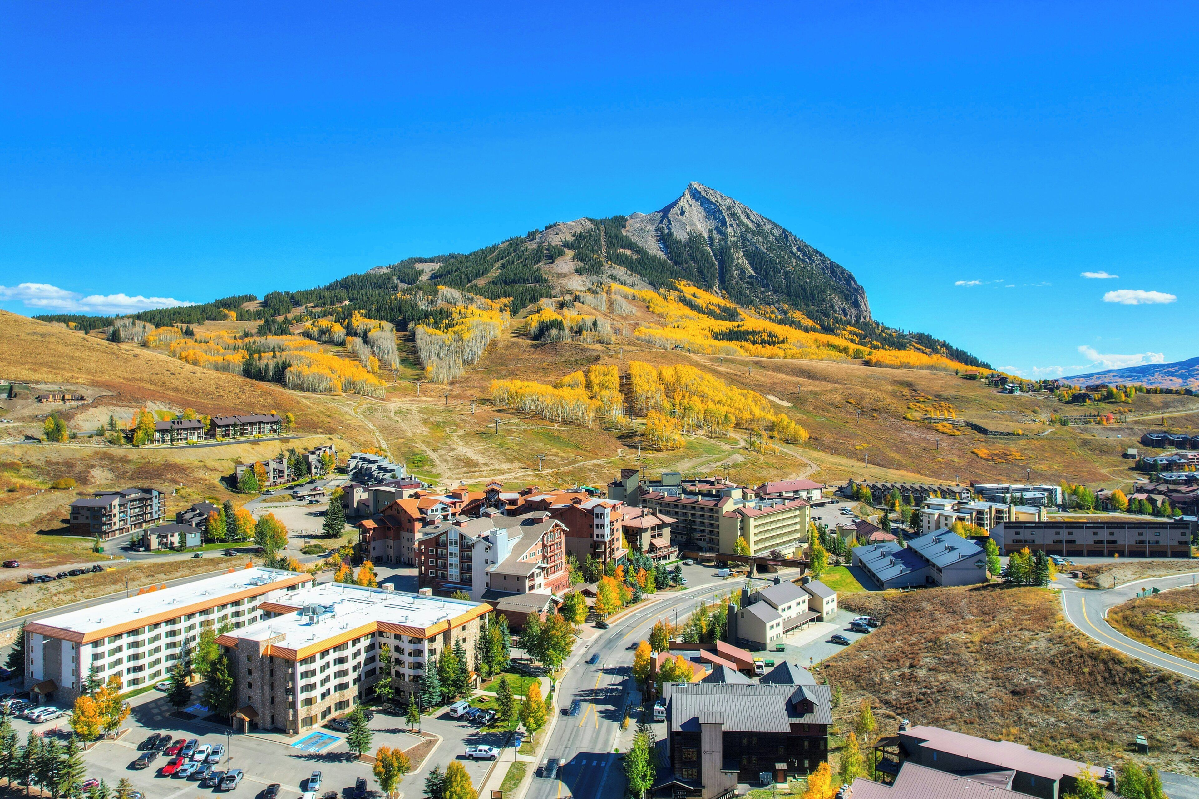 A town with a prominent mountain in the background, featuring a mix of modern and traditional buildings, a central road, and a parking area.