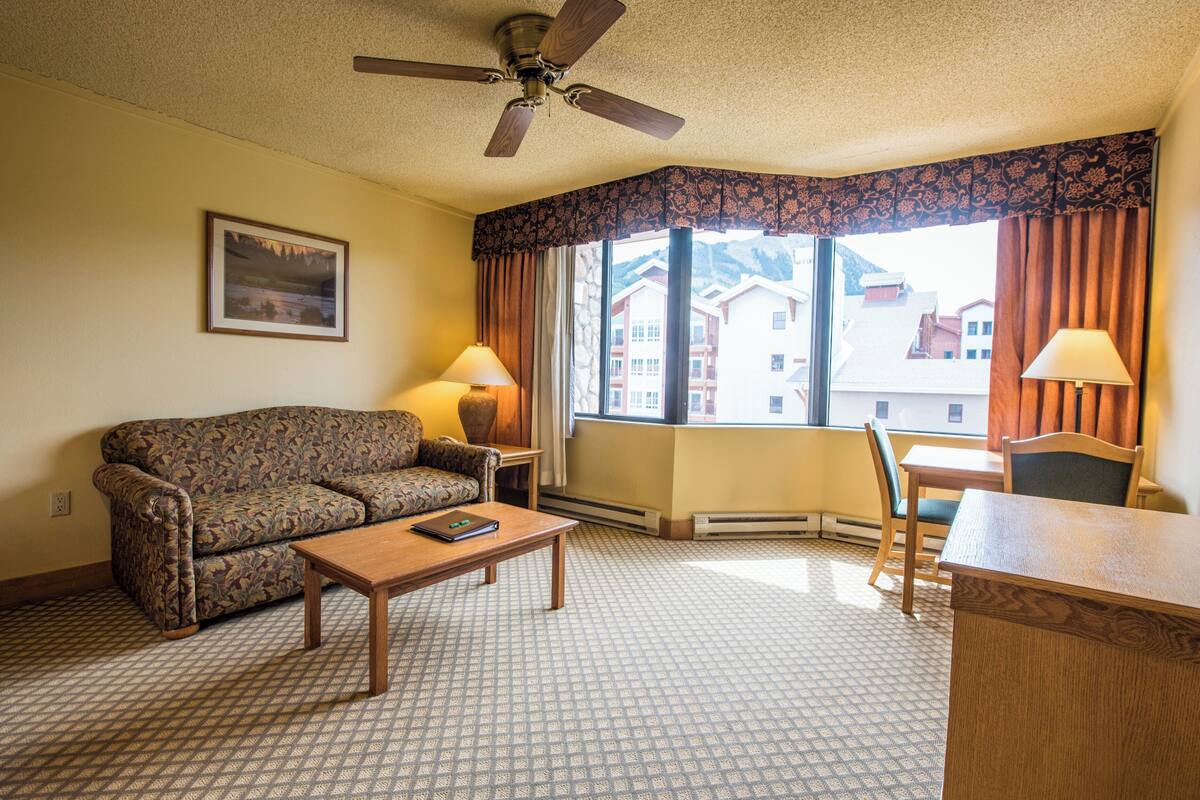 A hotel room with a patterned sofa, a wooden coffee table, a desk, two lamps, a ceiling fan, and a large window with curtains.