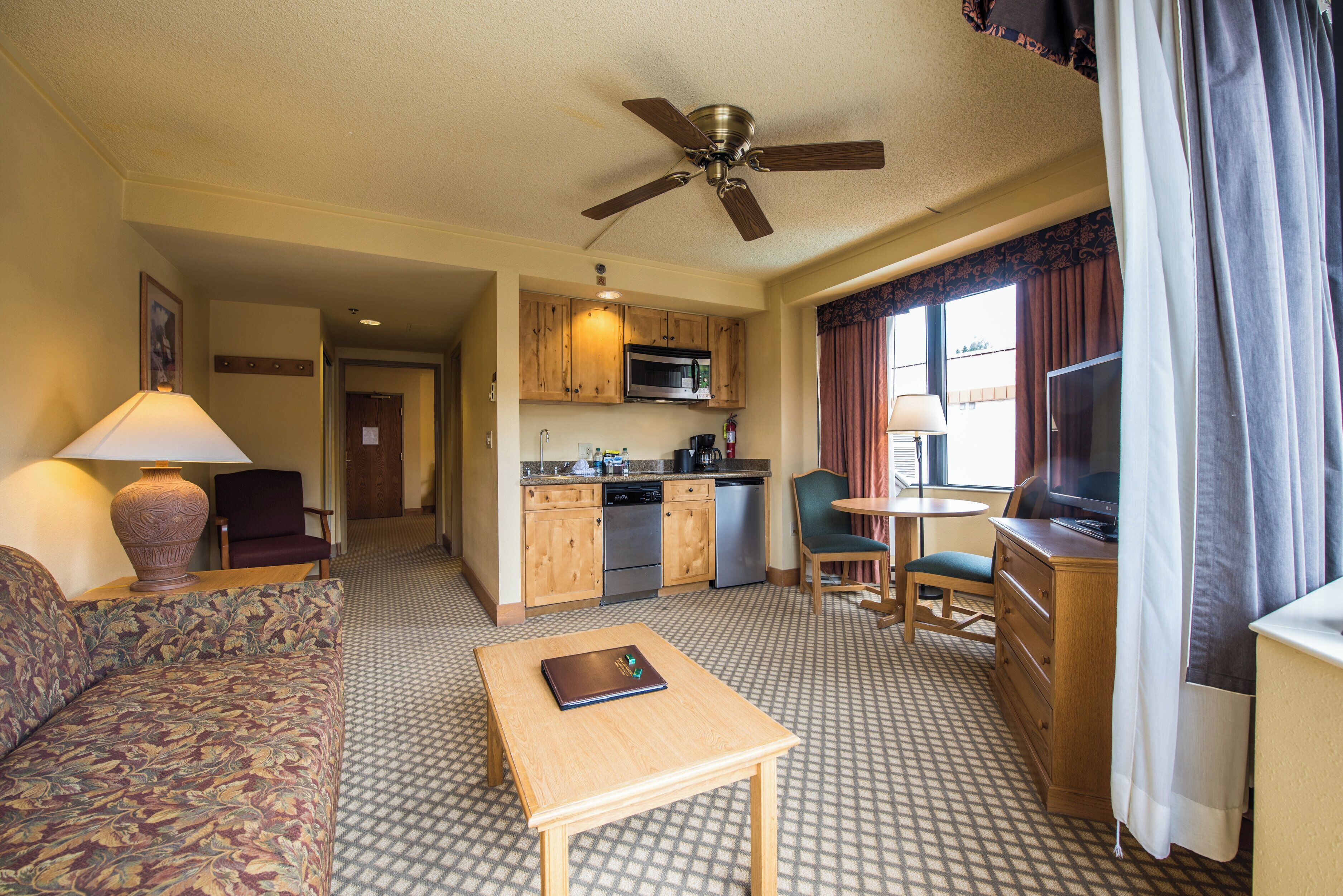 Quarto de hotel com cozinha compacta, sofá, mesa, TV e ventoinha no teto.