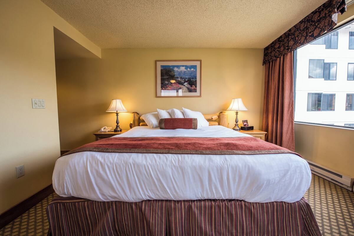 A hotel room with a large bed, two bedside lamps, a nightstand with a phone, a framed picture on the wall, and a window with curtains.