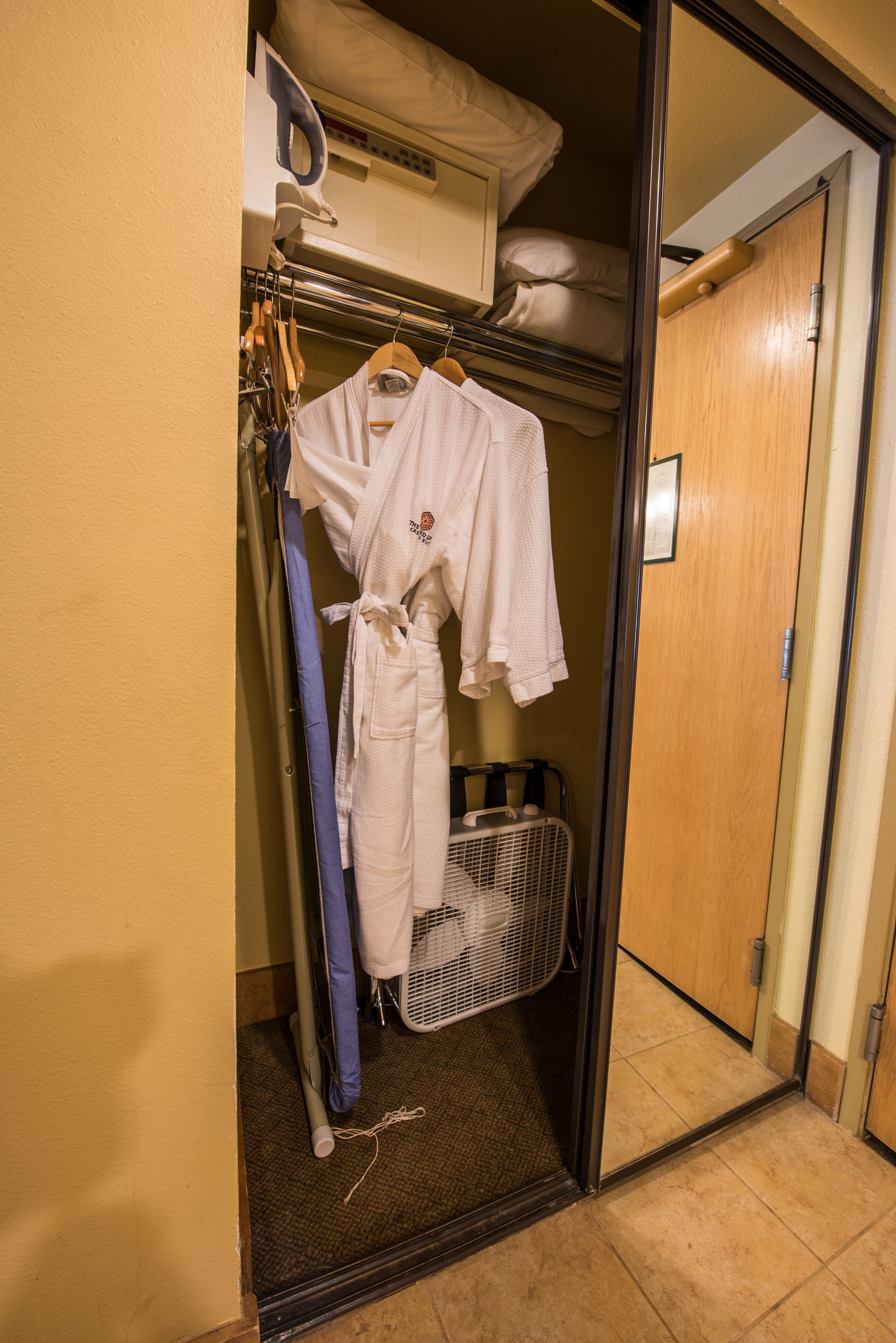A hotel room with a walk-in closet containing a hanging rack with towels and robes, a fan, and a suitcase.