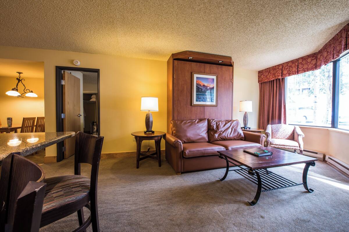 A hotel room with a dining area, a brown leather sofa, a coffee table, and a patterned armchair.