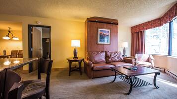 A hotel room with a dining area, a brown leather sofa, a coffee table, and a patterned armchair.