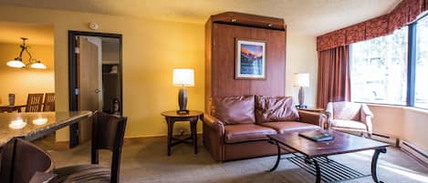 A hotel room with a dining area, a brown leather sofa, a coffee table, and a patterned armchair.