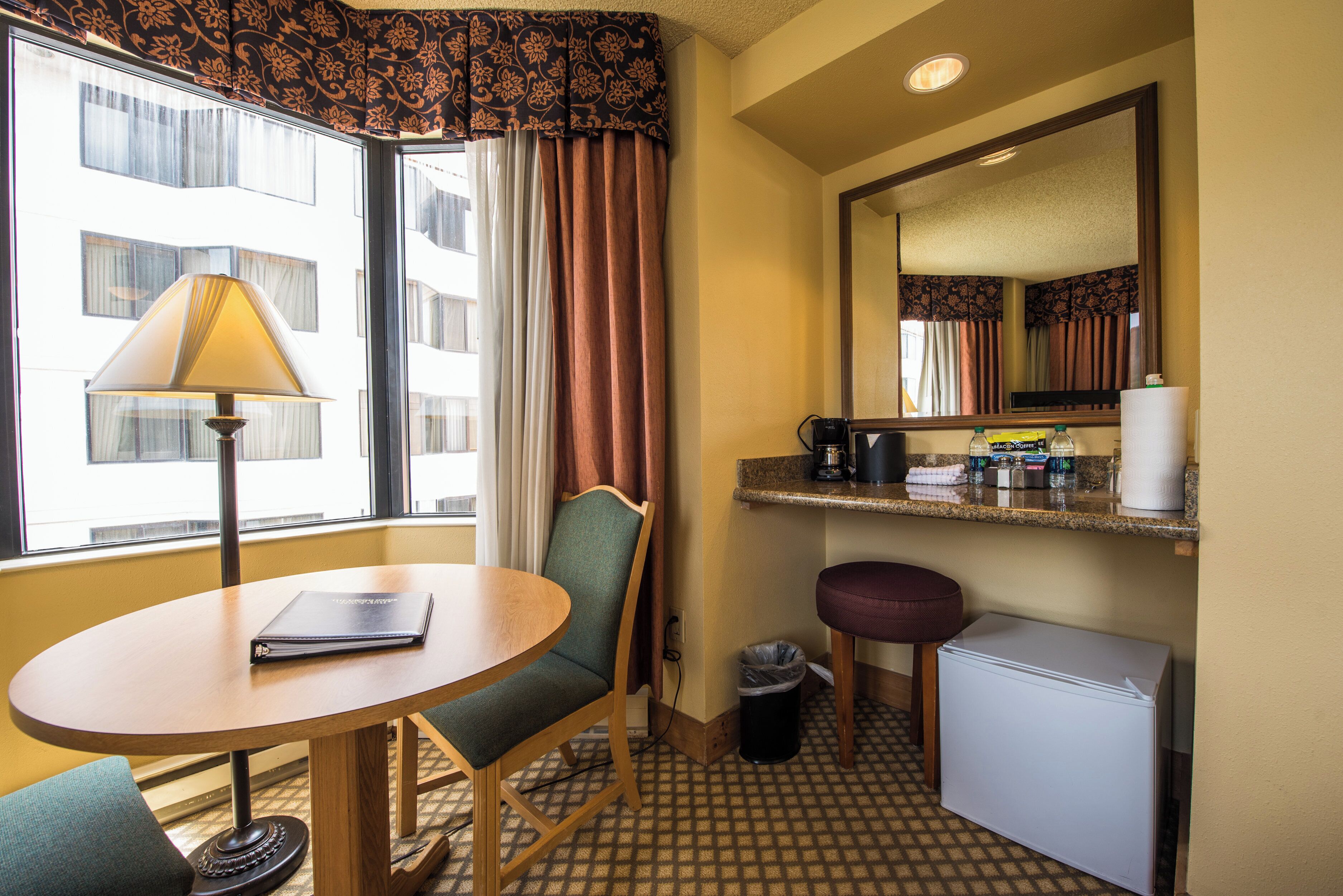 A compact hotel room with a small round table, a chair, a desk, a mirror, a mini-fridge, a trash bin, and a window with curtains.