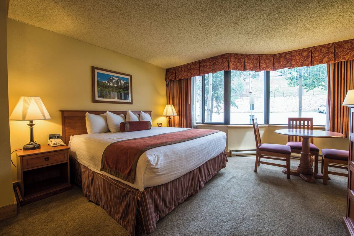 A hotel room with a large bed, a bedside table with a lamp, a small table with chairs, and a window with curtains.