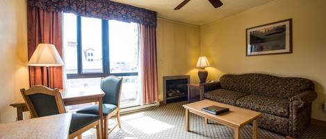A hotel room with a dining table, two chairs, a sofa, a coffee table, a fireplace, a ceiling fan, and a window with curtains.
