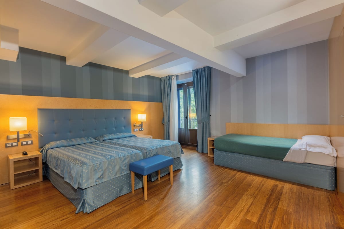 A hotel room with a large bed, a smaller bed, a bench, and a nightstand with a lamp.