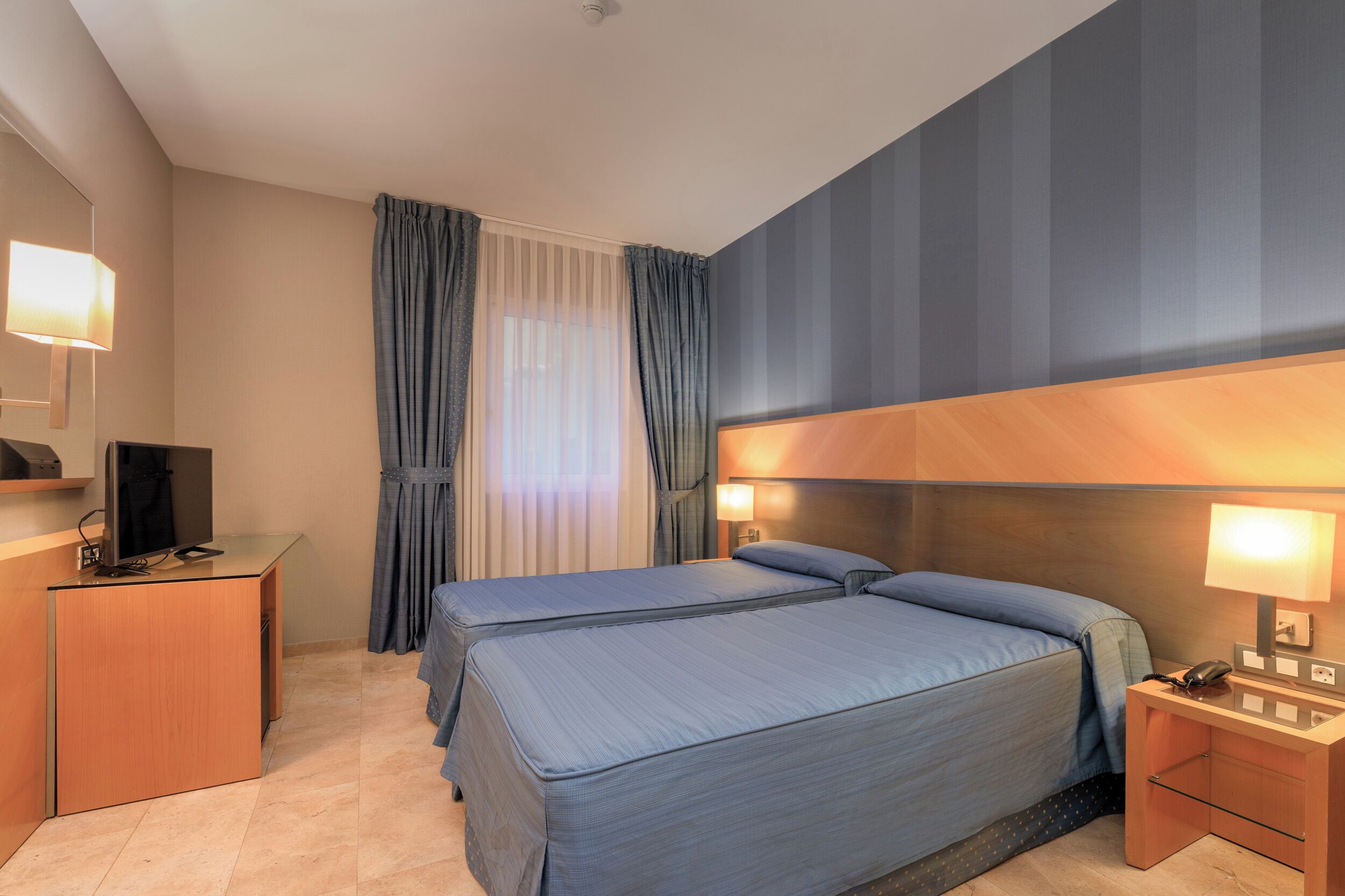 Une chambre d’hôtel avec un lit, des tables de chevet, un bureau avec un ordinateur et une fenêtre avec des rideaux.