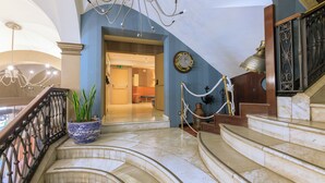 A grand staircase with marble steps leading to a second floor, a potted plant, a chandelier, and a clock on the wall. - Hotel Del Mar (Barcelona)