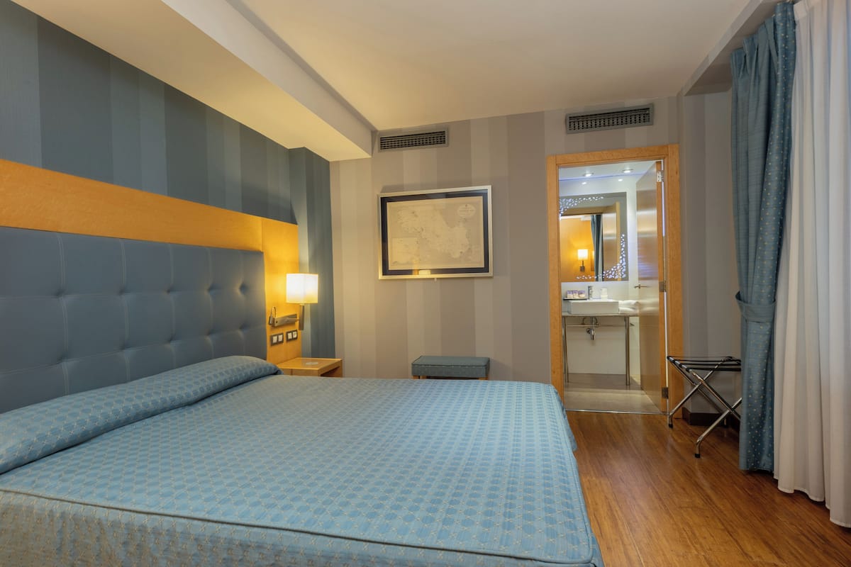 A hotel room with a large bed, a bedside table with a lamp, a framed map on the wall, a bathroom with a mirror and sink, and a drying rack.