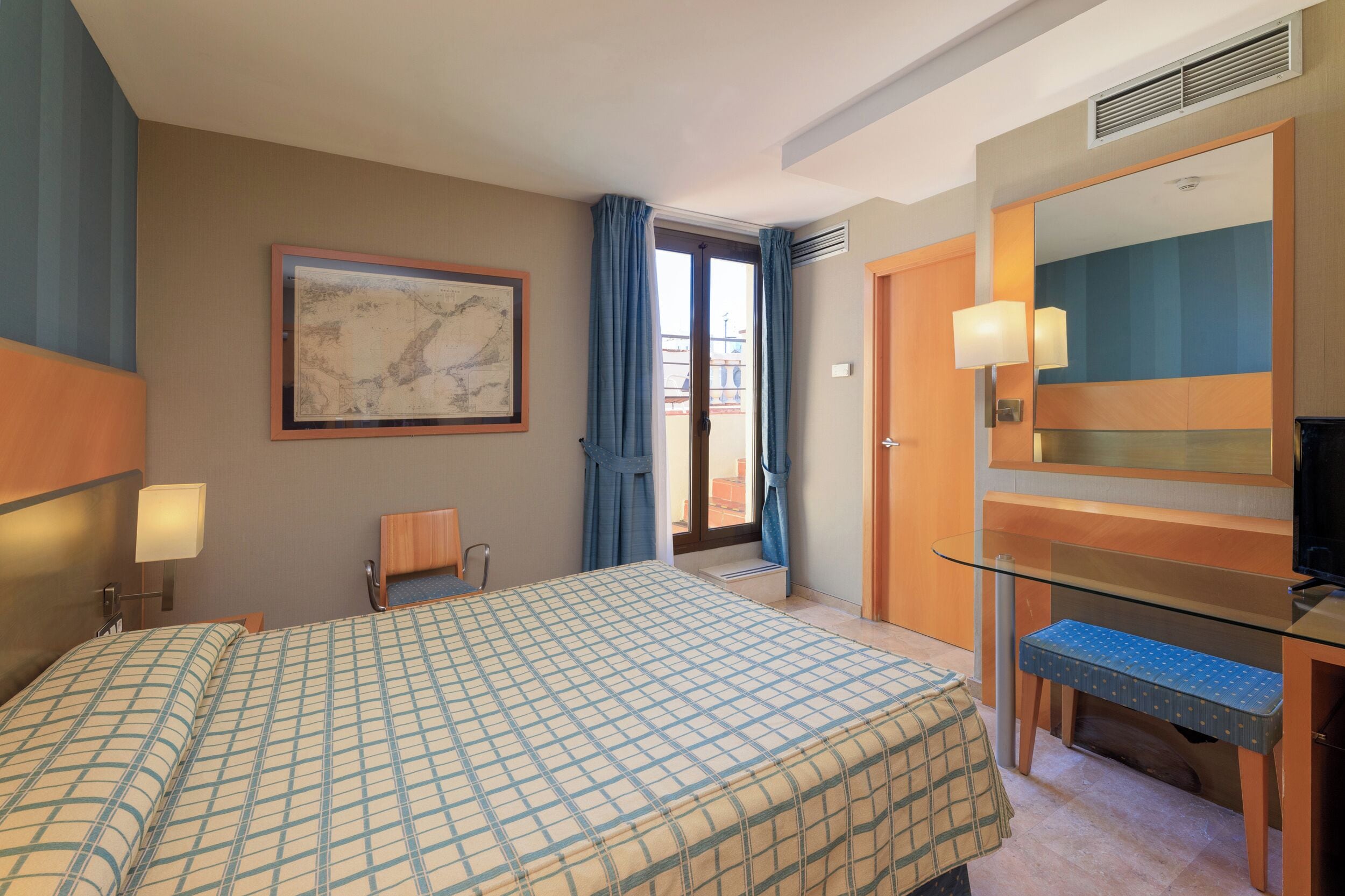 Habitación de hotel con cama, escritorio, silla, televisor y un amplio ventanal con cortinas.