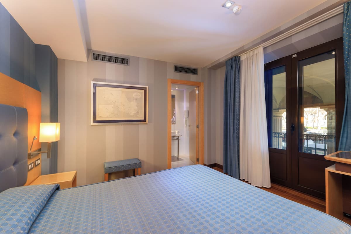 A hotel room with a large bed, a bedside table, a lamp, a bench, a framed artwork on the wall, and a view through a door to the outside.