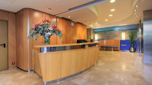 A modern reception area with a curved wooden reception desk, a large floral arrangement, a reception desk with a computer, a seating area, and a mural on the wall. - Hotel Del Mar (Barcelona)