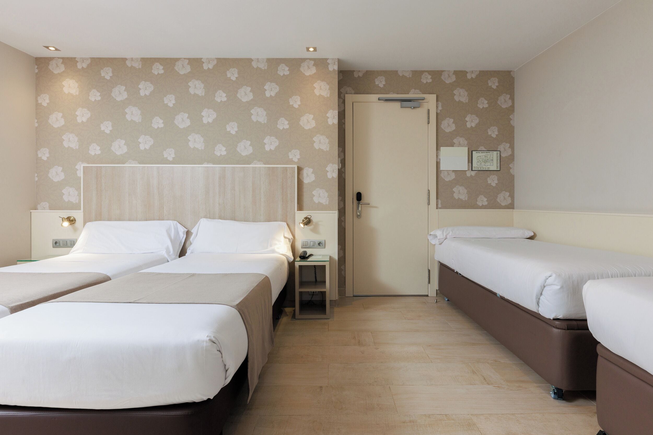 A hotel room with two beds, a small bedside table, a nightstand, a door, and a wall-mounted light fixture.