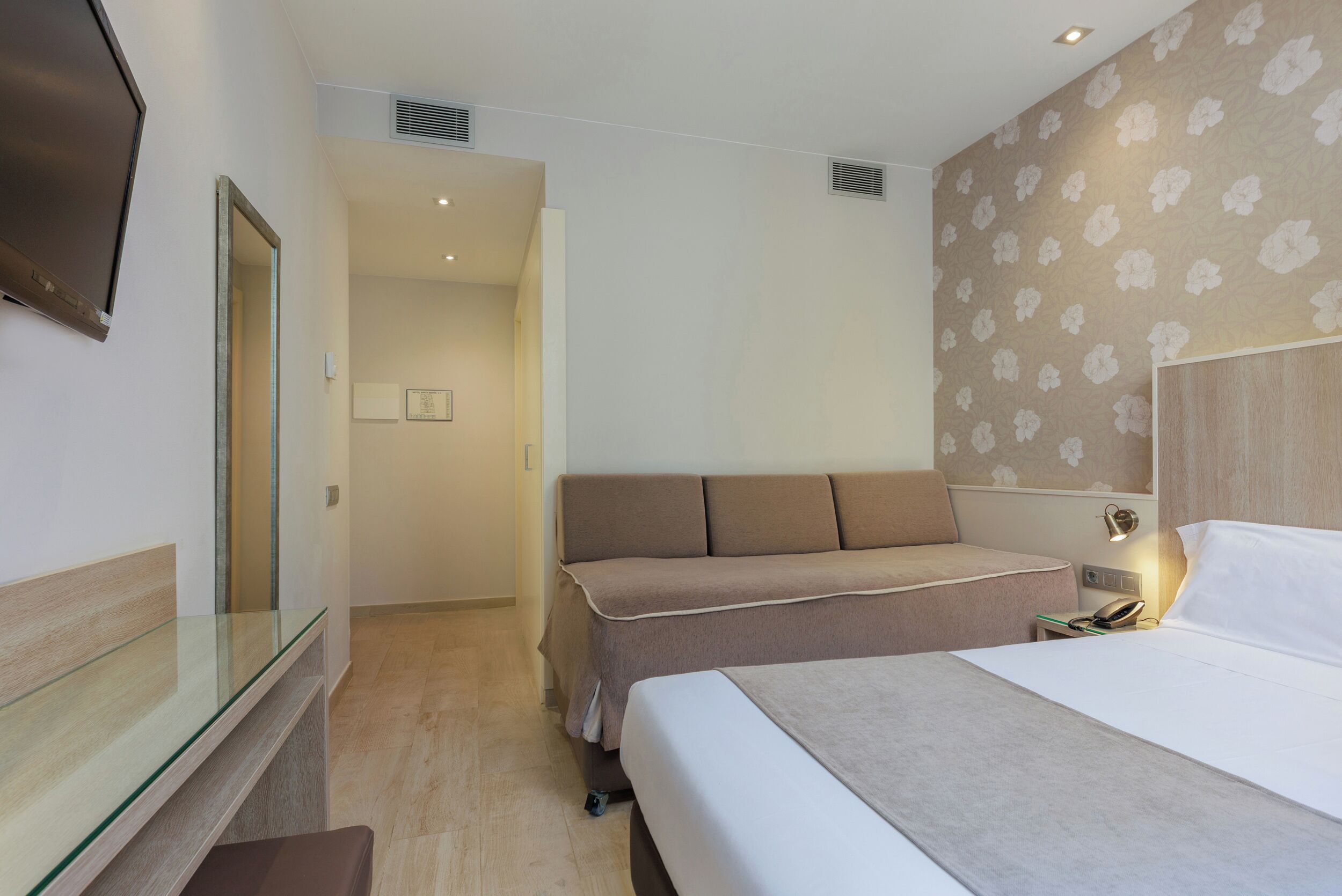 A hotel room with a bed, a sofa, a glass partition, a bedside table, and a wall-mounted TV.