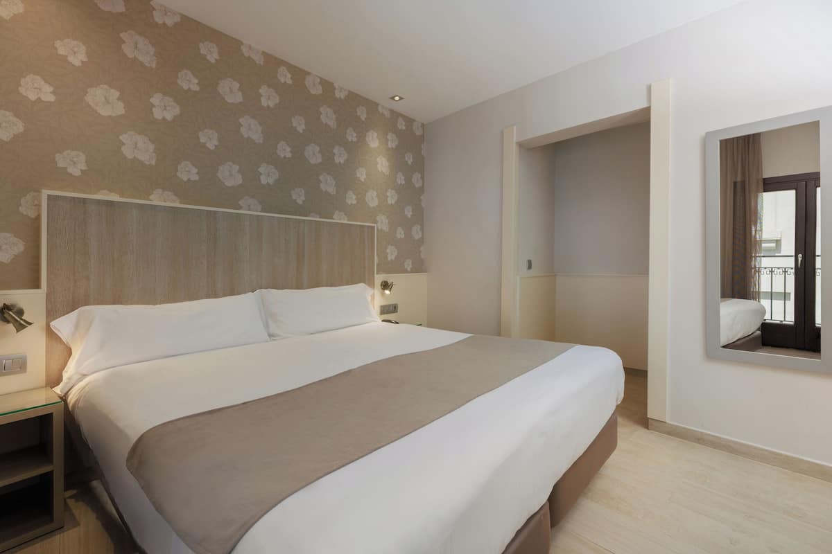 A modern hotel room with a large bed, a nightstand, a mirror, and a wall with a floral pattern.