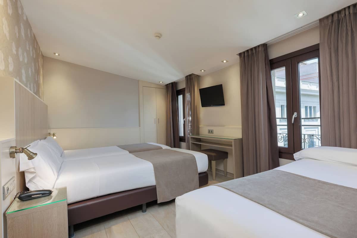 A hotel room with two beds, a desk, a flat-screen TV, and a window with curtains.