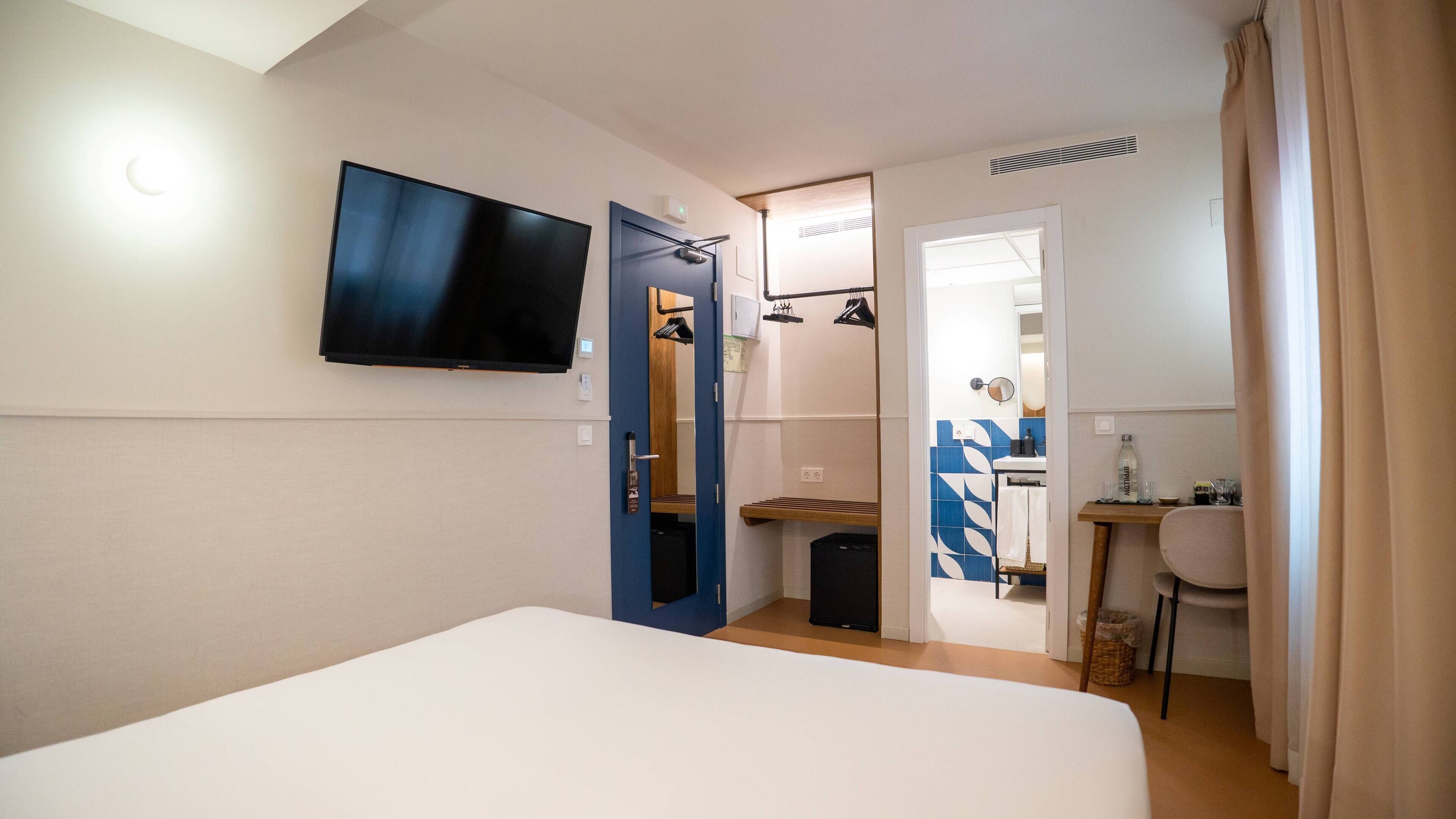 A hotel room with a bed, a TV mounted on the wall, a mirror, a desk with a chair, and a bathroom visible through an open door.