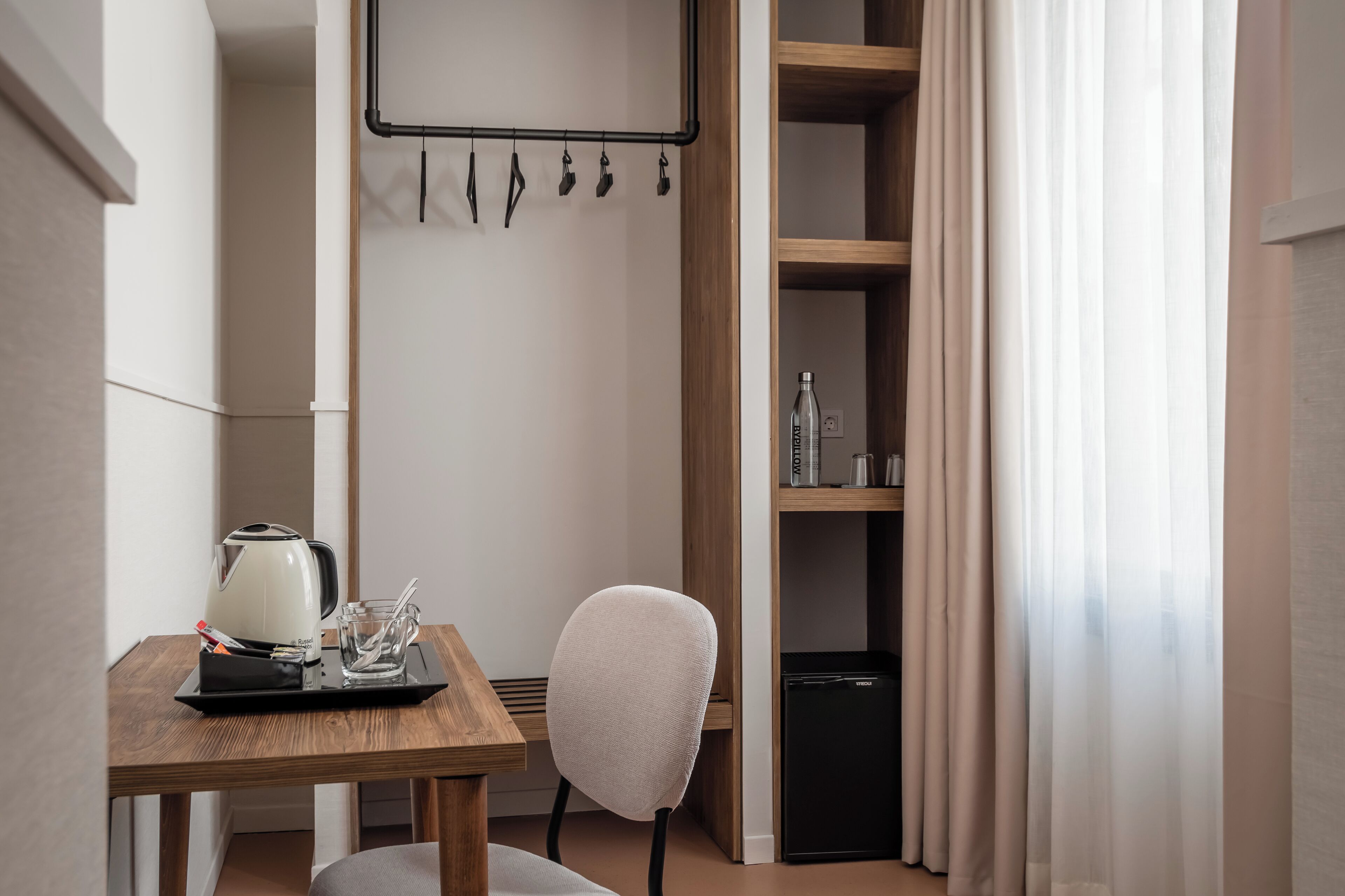 Una kitchenette pequeña con un hervidor, un vaso y una bandeja sobre una mesa de madera. Hay una silla y un refrigerador negro en la esquina.