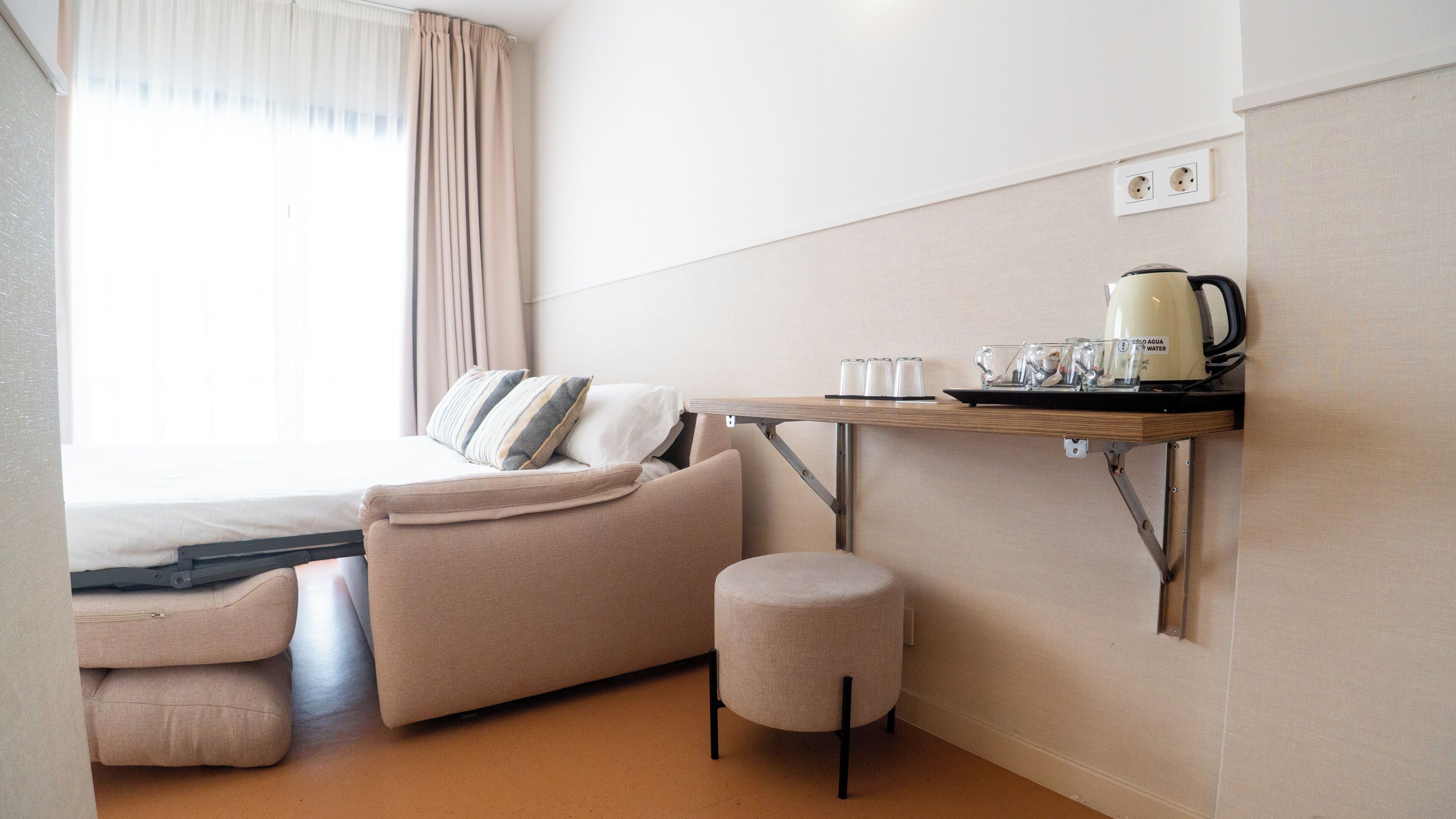 A modern hotel room with a sofa, ottoman, and a small table with a kettle and glasses.