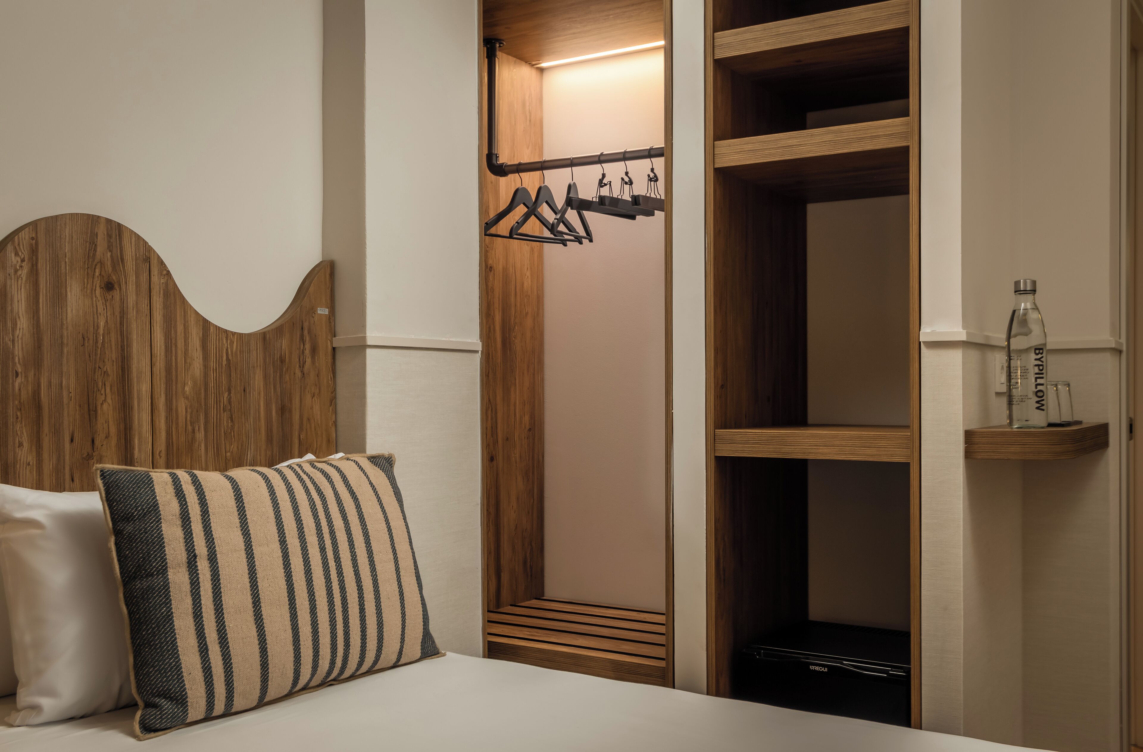 A bedroom with a wooden headboard, a striped pillow, a wooden closet with a hanging rack, and a shelf with bottles.