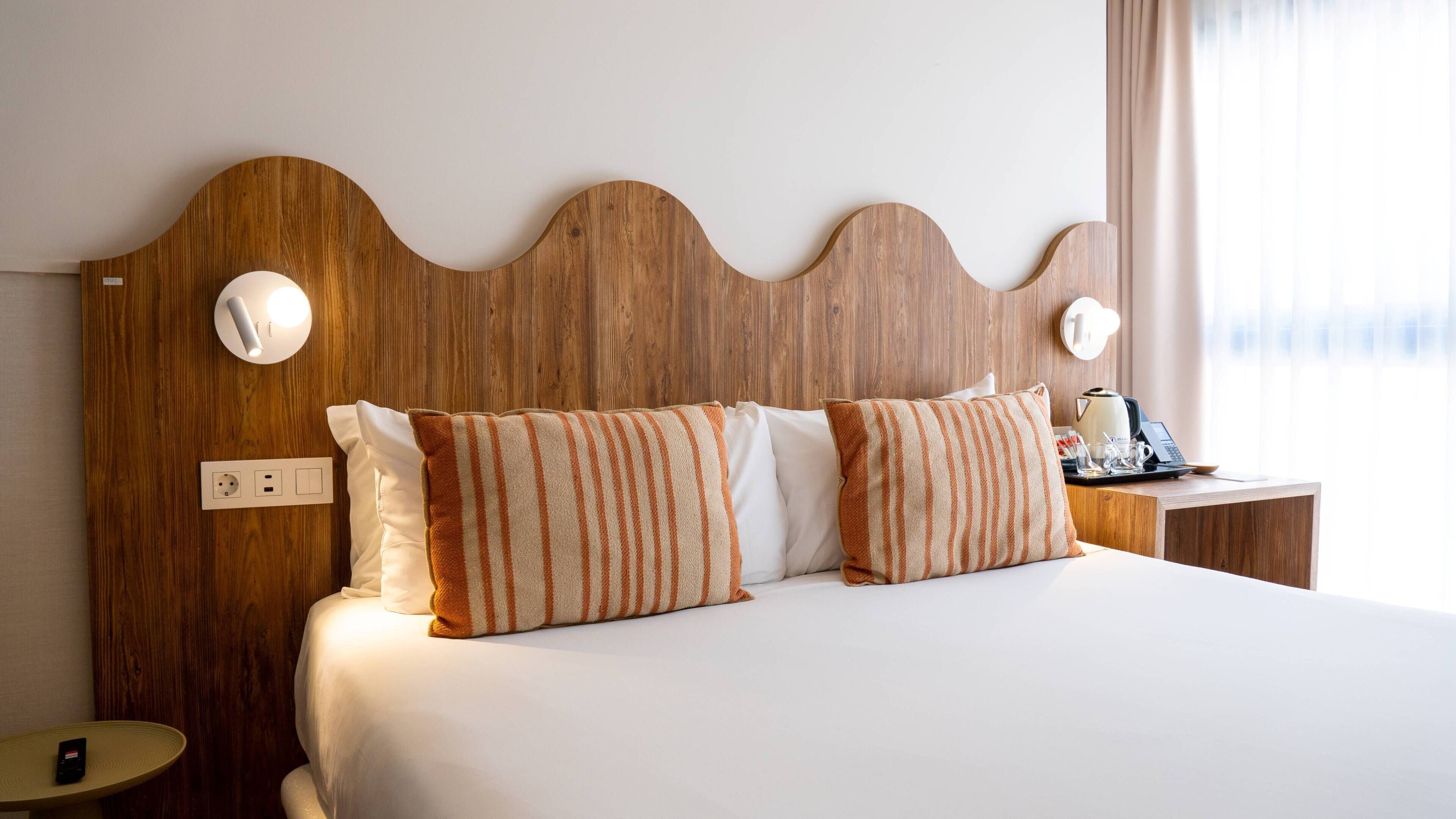 A hotel room with a wooden headboard, a bed with white bedding, striped pillows, a bedside table with a kettle, and a lamp.