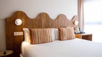 A hotel room with a wooden headboard, a bed with white bedding, striped pillows, a bedside table with a kettle, and a lamp.