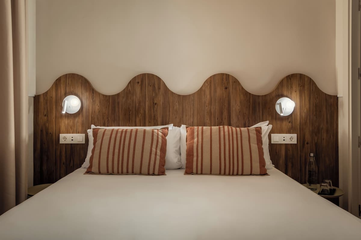 A neatly made bed with striped pillows, a wooden headboard with integrated lighting, and a bedside table with a bottle and a cup.