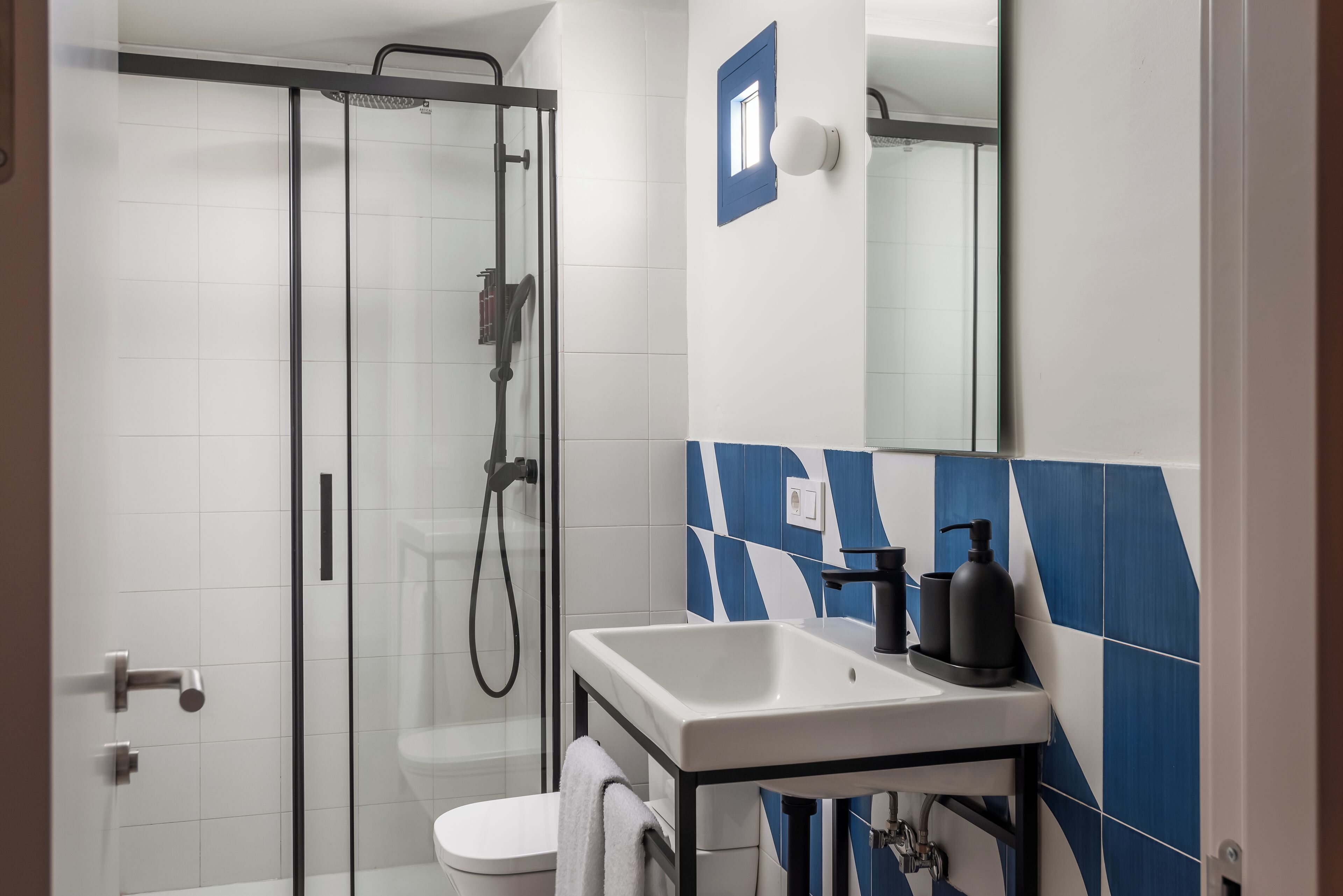 Une salle de bain moderne avec une douche à parois de verre, des carreaux blancs, un lavabo blanc et des carreaux bleus à motifs géométriques sur le mur.