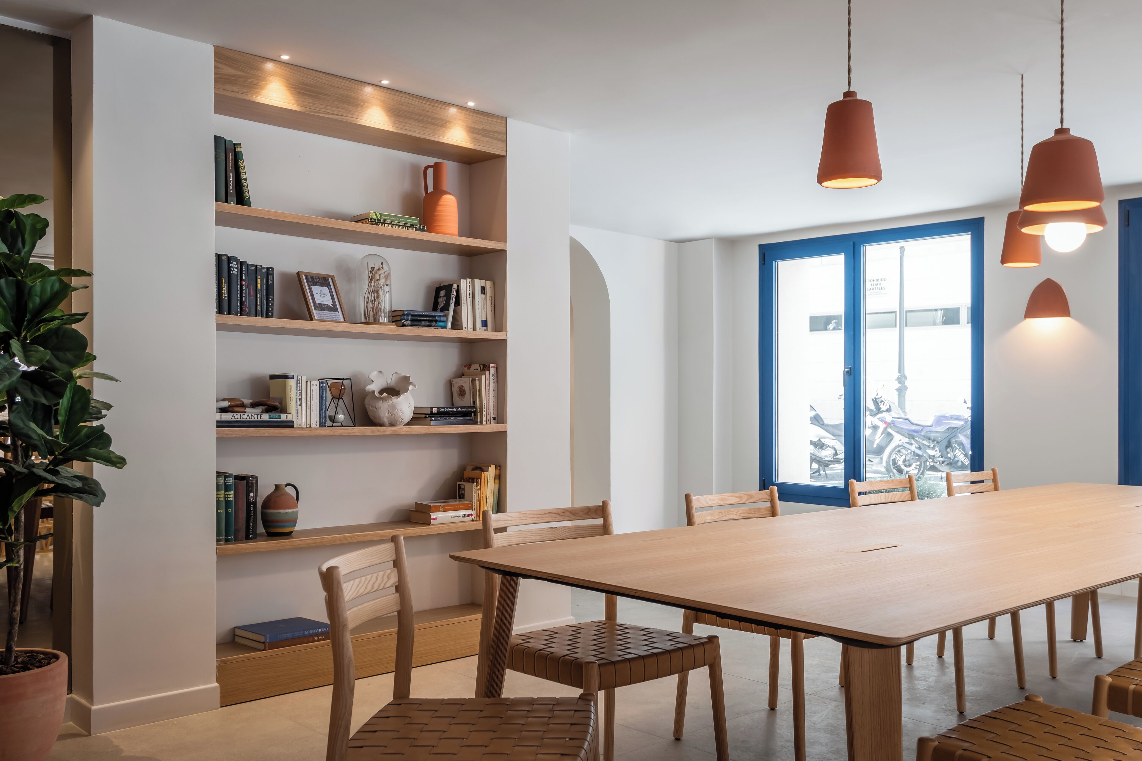 A modern dining area with a wooden table and chairs, a bookshelf with books and decorative items, and pendant lights.