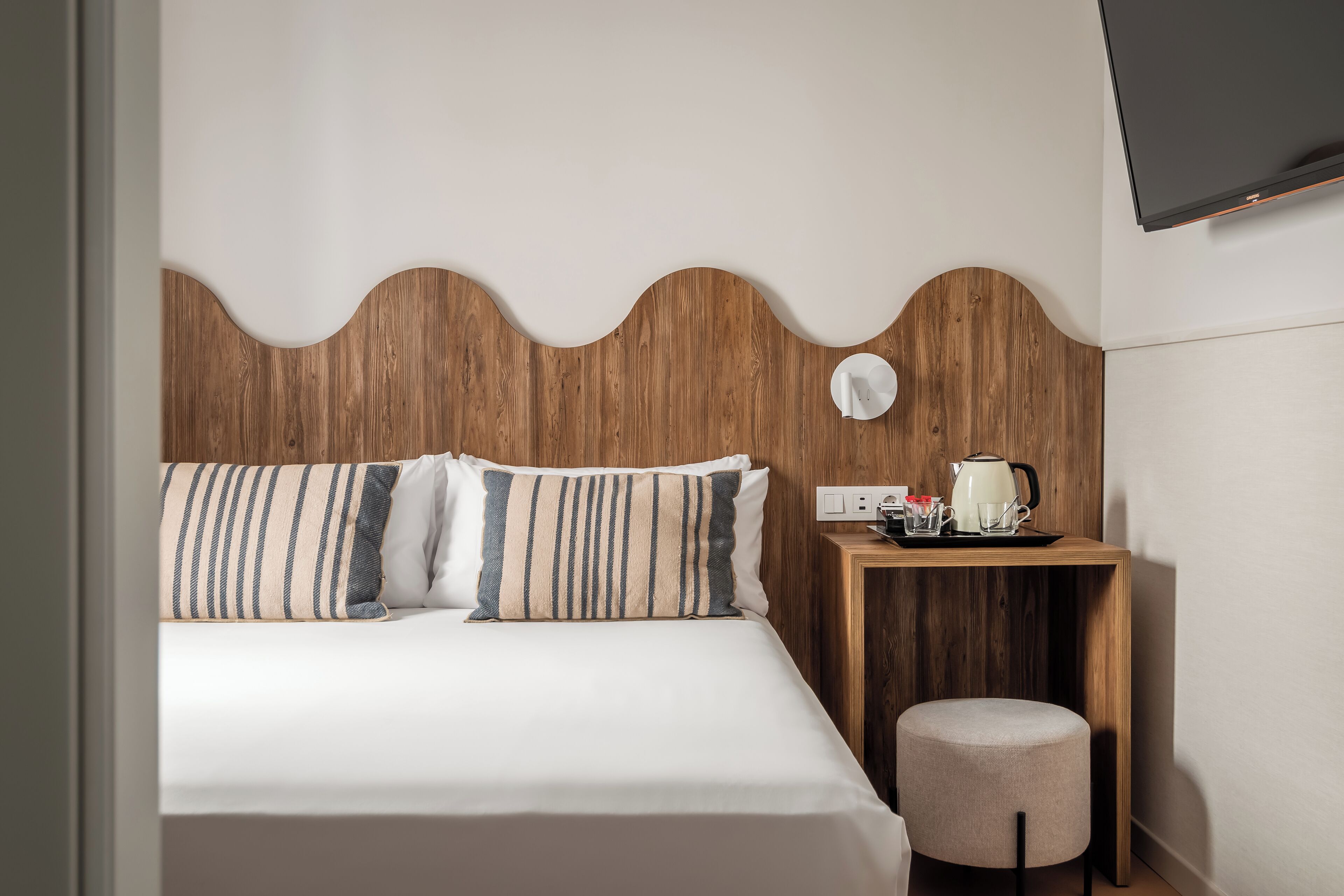 A neatly made bed with striped pillows, a wooden headboard, a bedside table with a kettle, and a small stool.