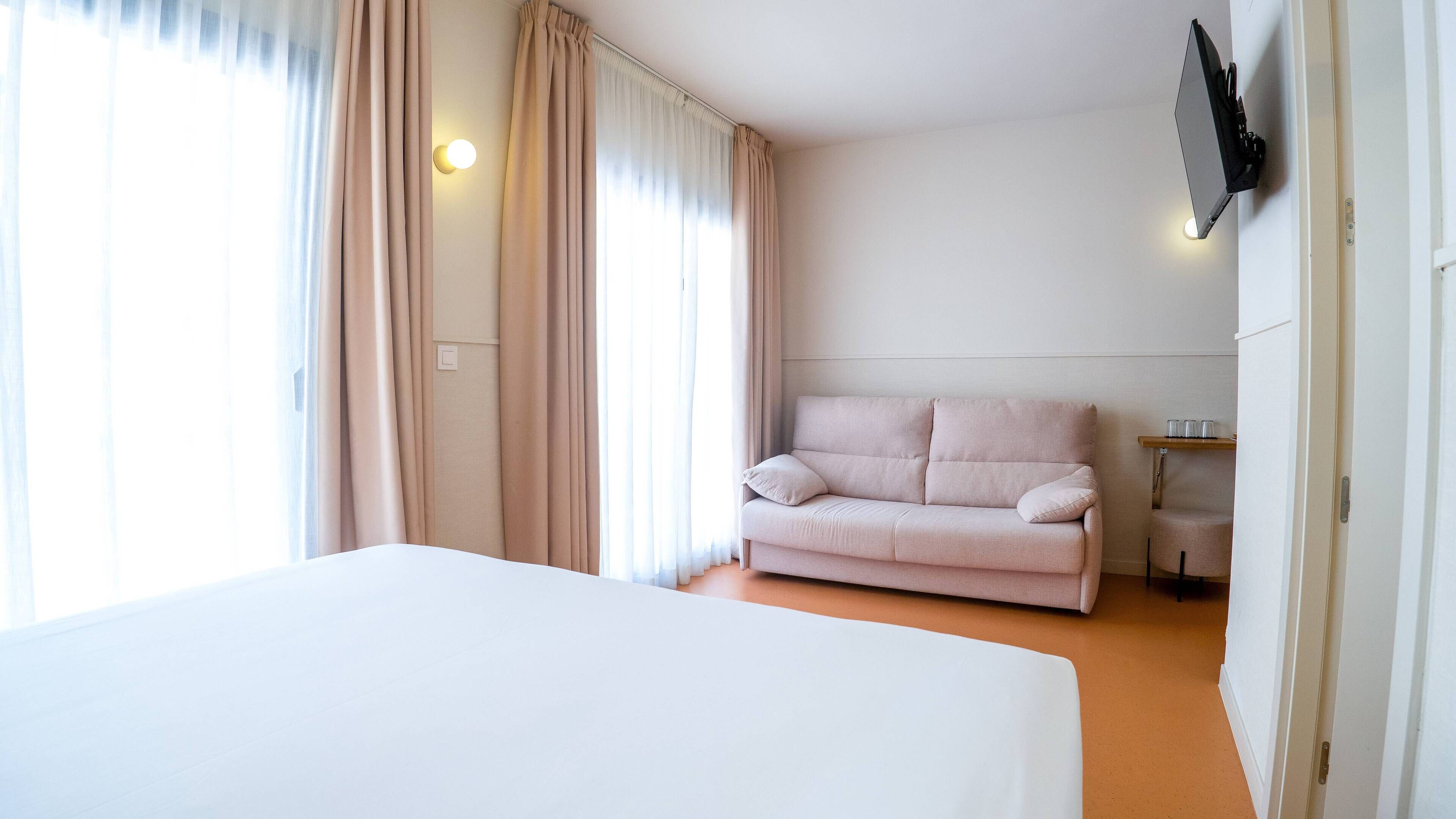 A hotel room with a bed, a sofa, a TV mounted on the wall, and a window with curtains.