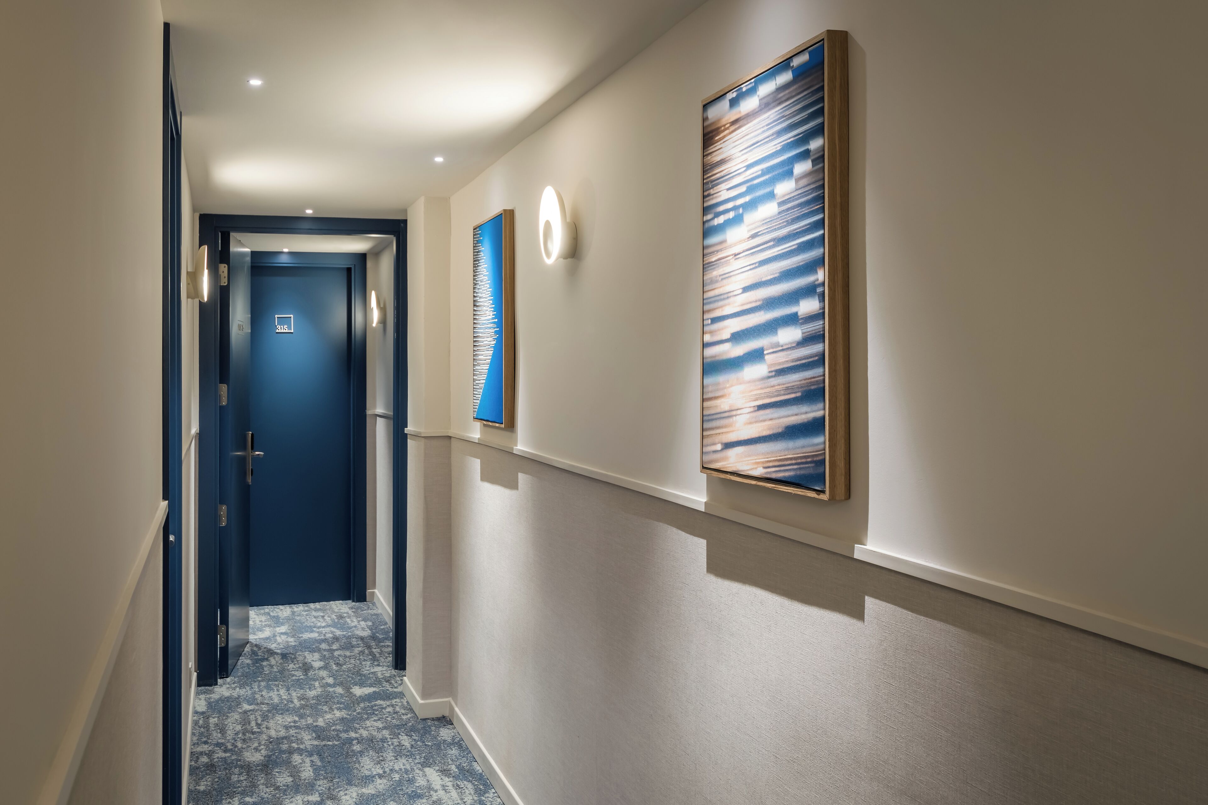 Un pasillo con una puerta azul al final, un interruptor de luz y dos cuadros en la pared.