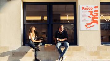 Dos personas sentadas en un alero, fuera de un café, con un cartel que muestra opciones de comida y bebida.