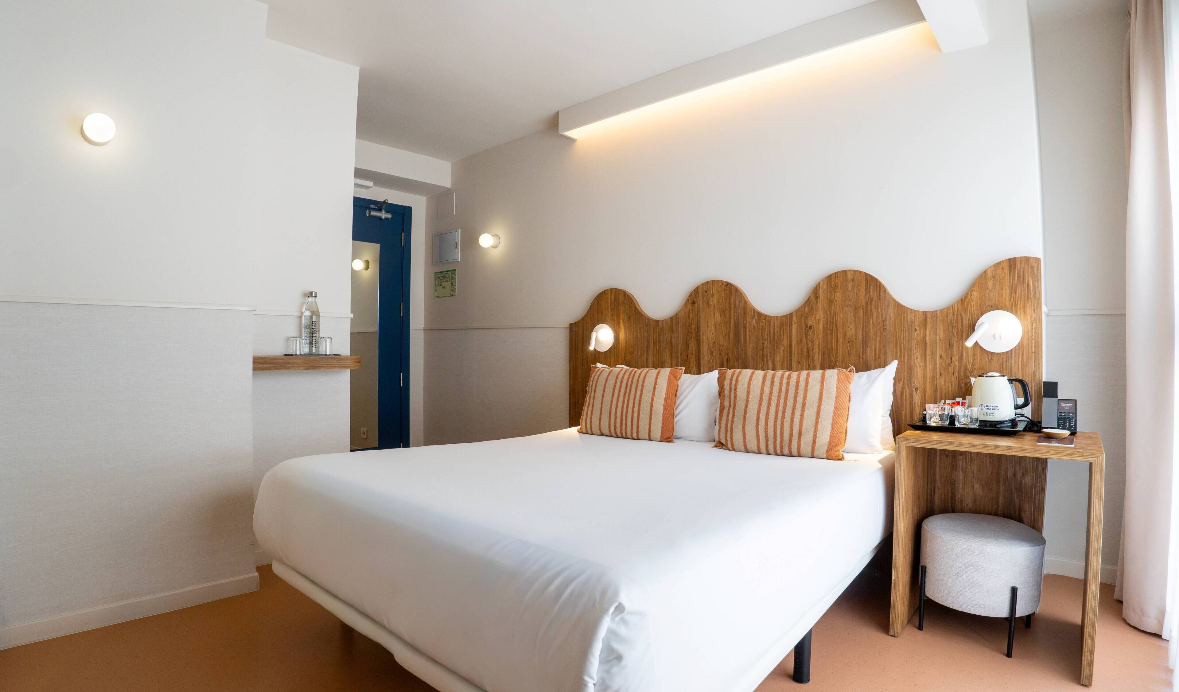 A hotel room with a large bed, a wooden headboard, bedside table, and a small desk with a kettle and coffee maker.