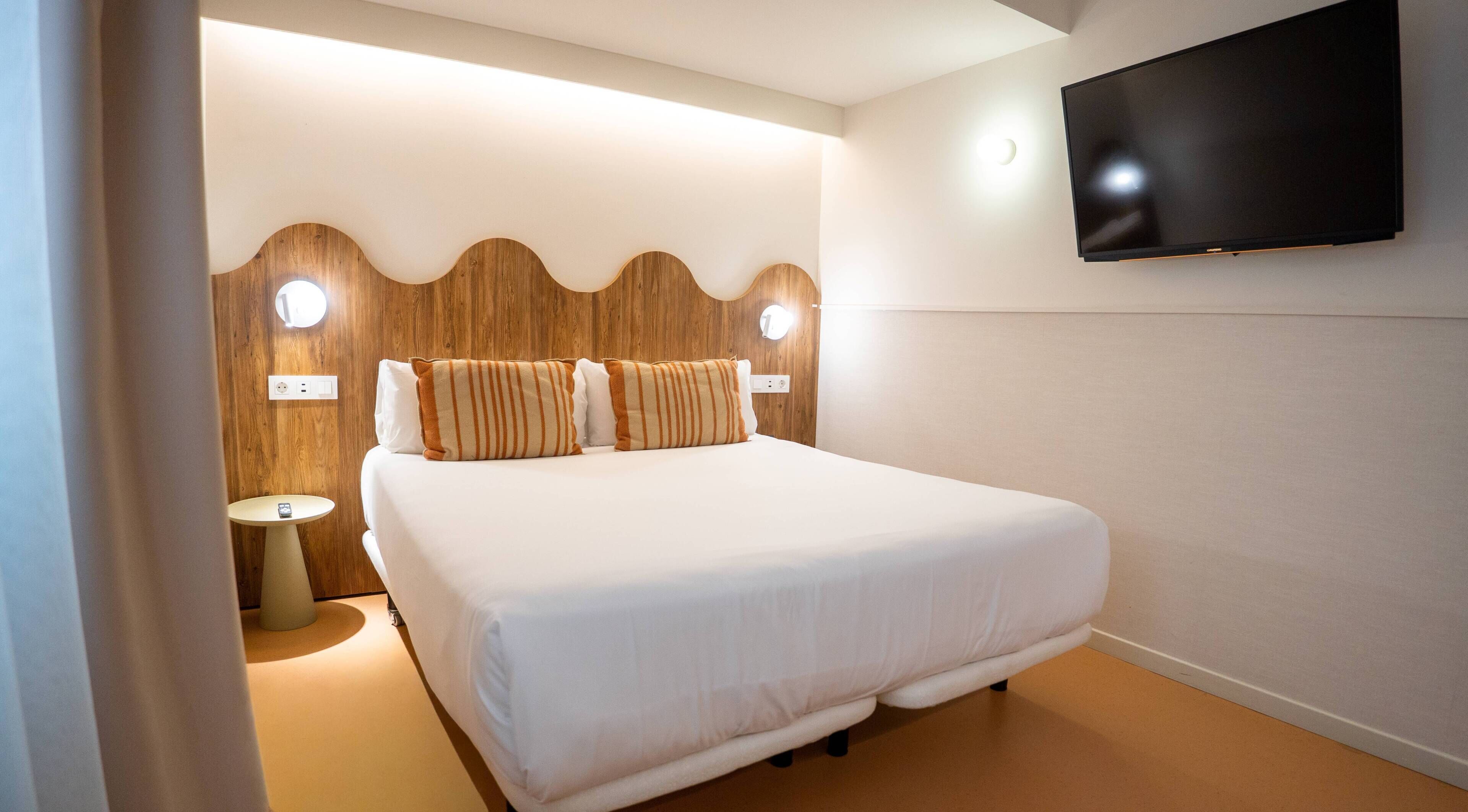Una habitación de hotel con una cama, un televisor montado en la pared y una mesita de noche redonda.