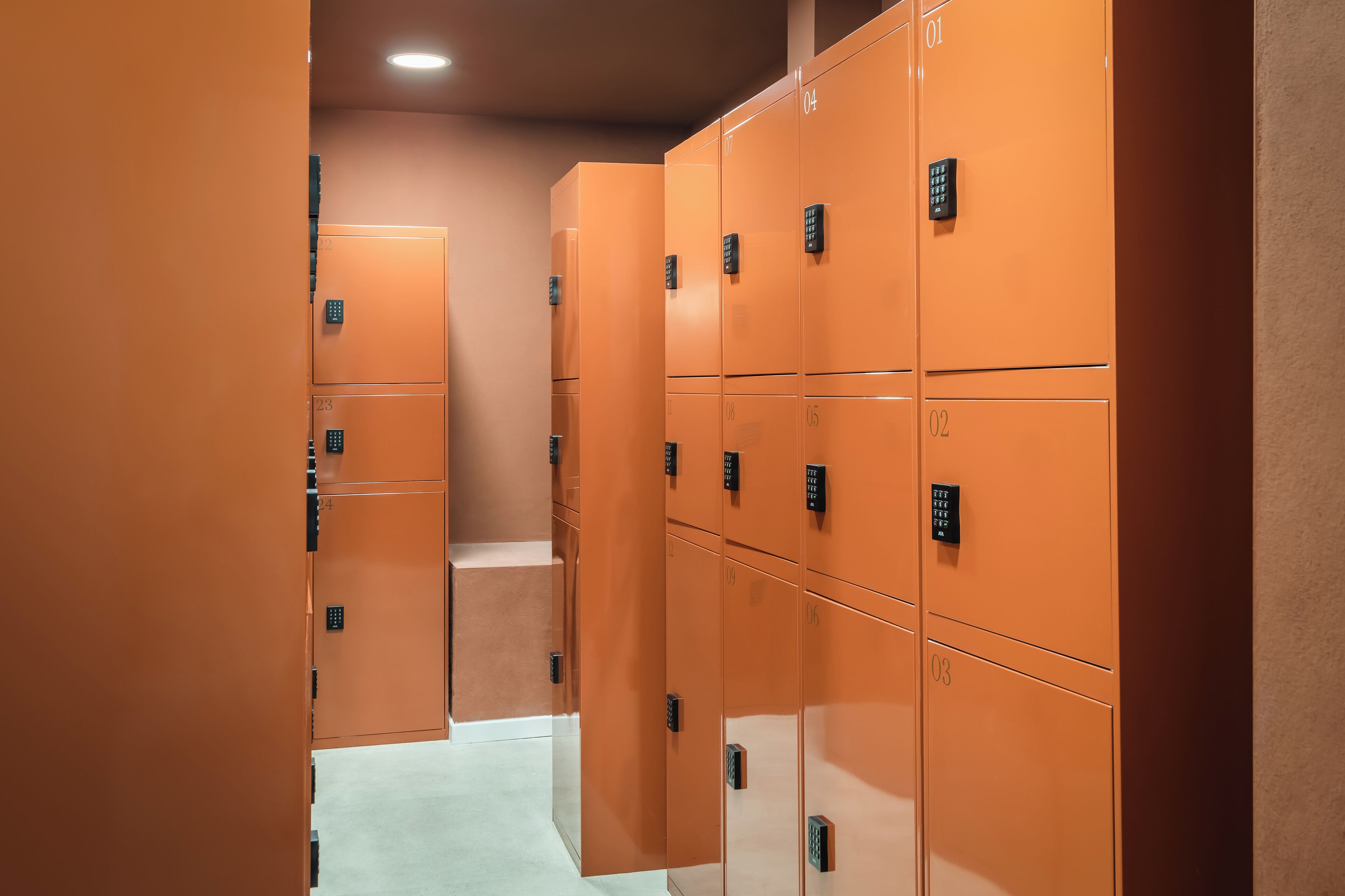Une rangée de casiers orange avec des portes numérotées, un sol de couleur claire et un luminaire encastré au plafond.