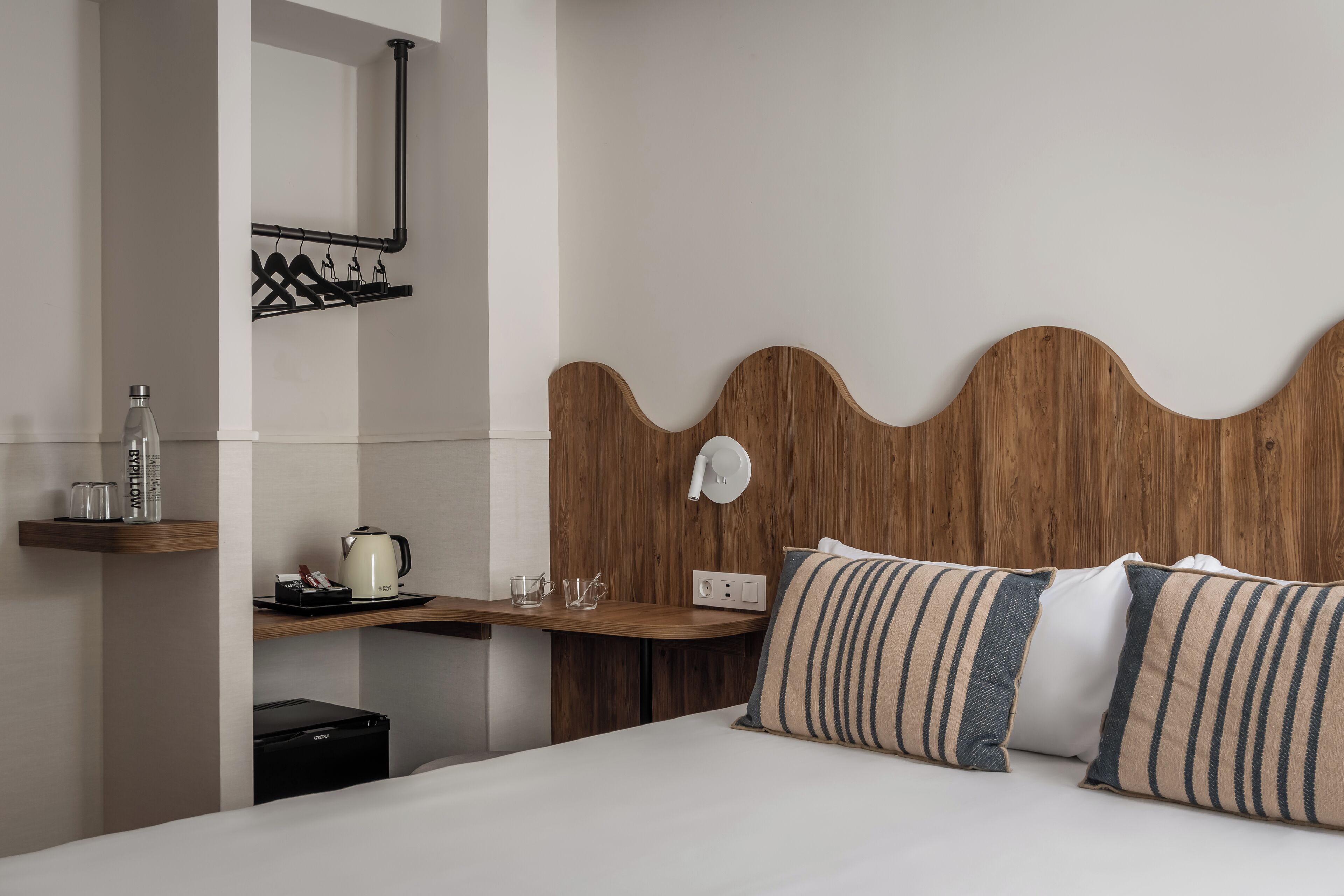 A hotel room with a wooden headboard, a bedside table with a kettle, glasses, and a bottle, and a bed with striped pillows.