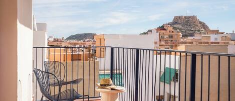 A balcony with a metal chair, a small table with a hat, and a view of buildings and a hill.