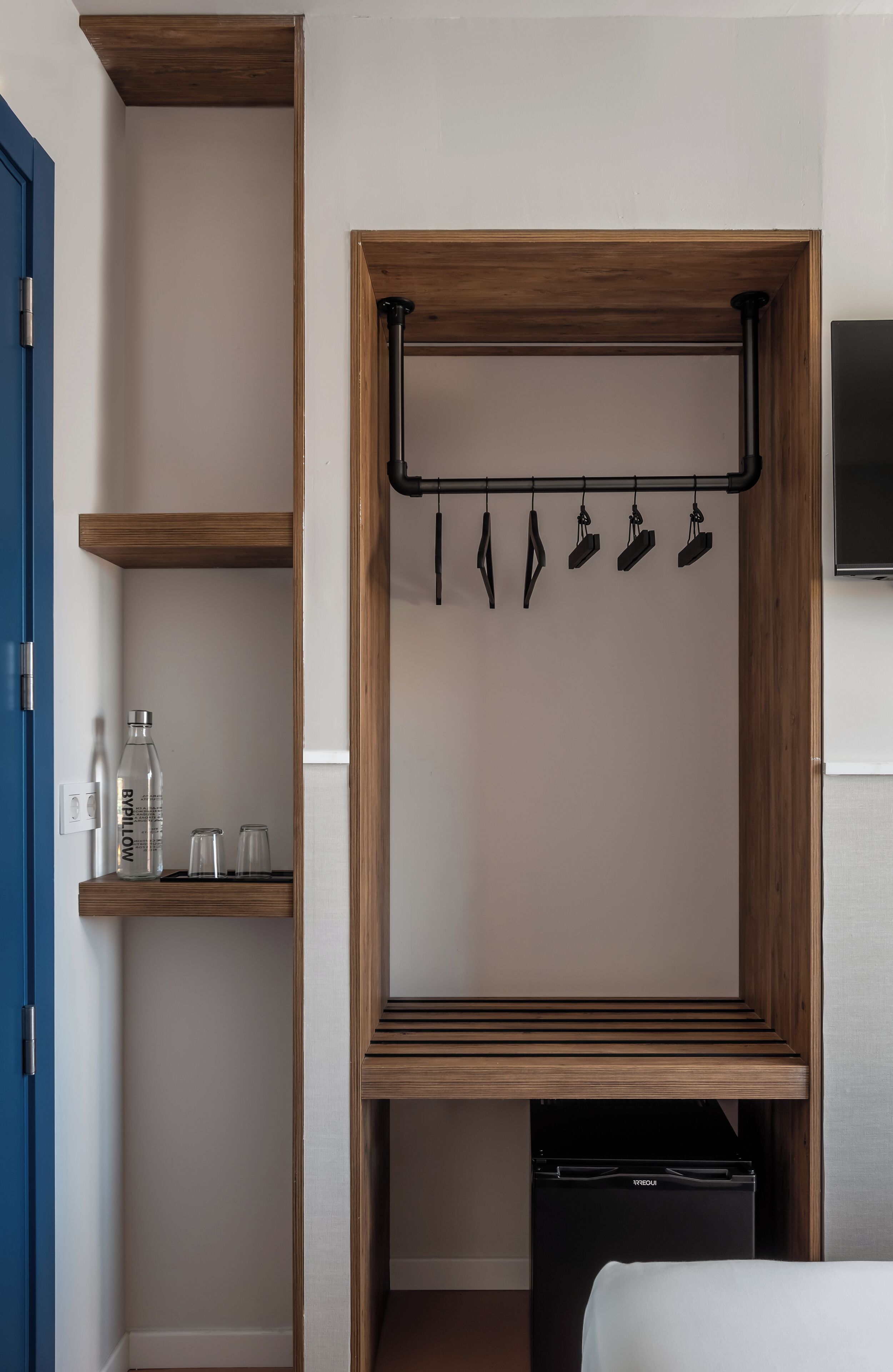 A compact room with a wooden wardrobe, a small shelf with bottles and glasses, a hanging rack with bags, and a black appliance.