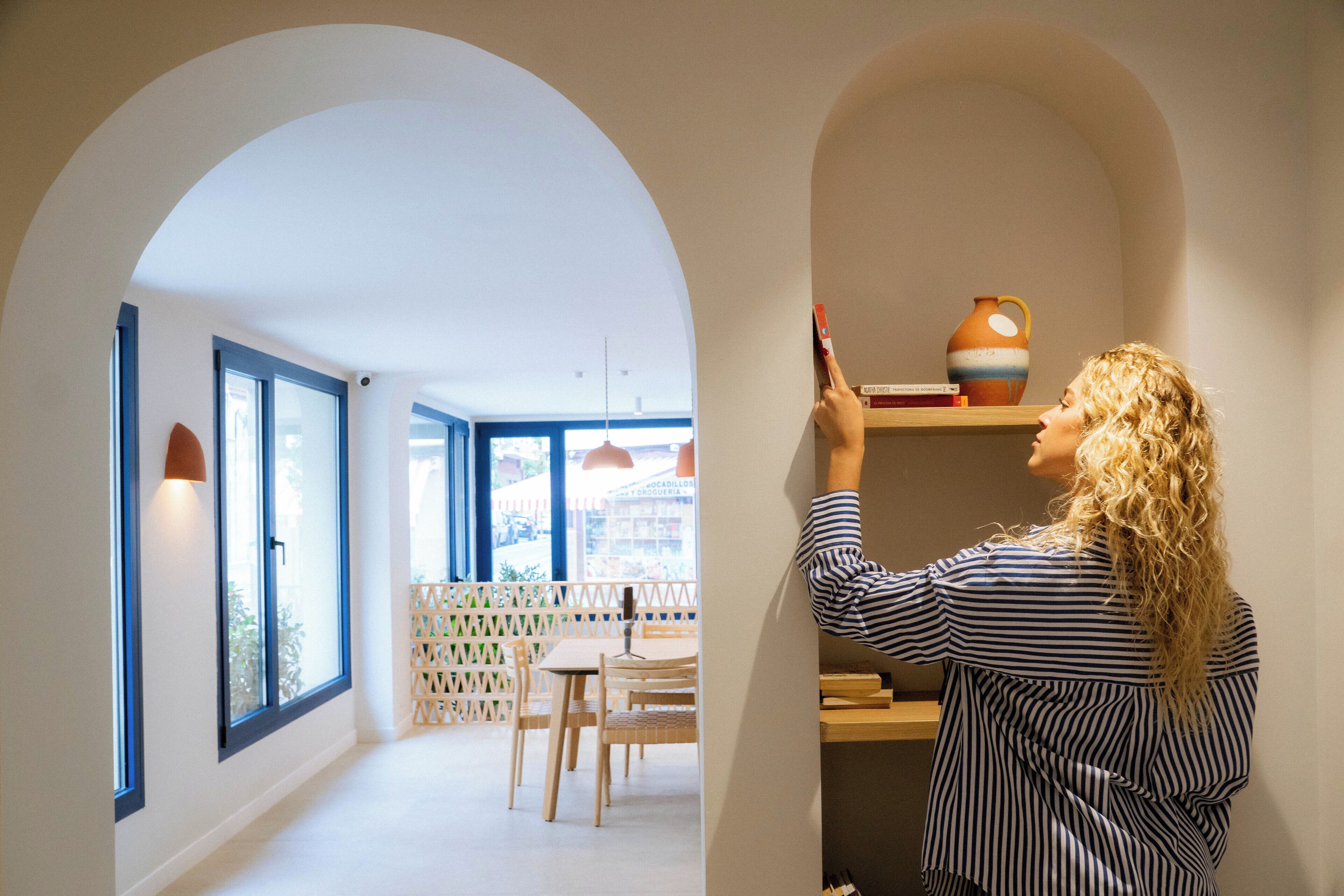 A person standing in a hallway with arched doorways, a dining area with wooden chairs and a table, and a shelf with books and a vase.