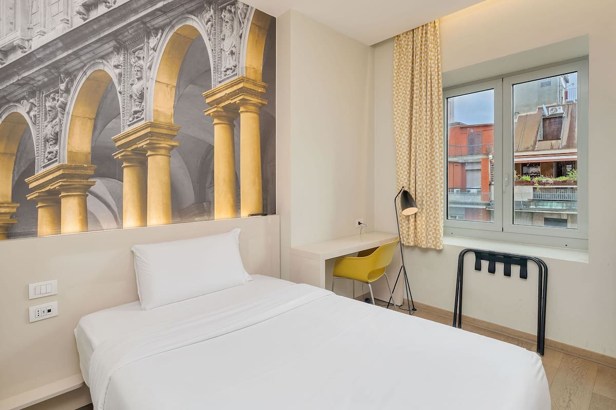 Et moderne hotelværelse med seng, skrivebord, stol, lampe og et stort vindue med udsigt til bygninger.